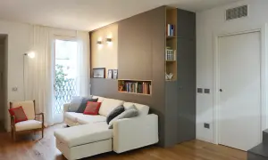 Divano bianco con cuscini grigi e rossi, accanto a una libreria a muro color un marrone chiaro.