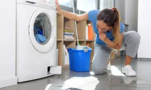 Donna preoccupata controlla la lavatrice che perde acqua da sotto. Sul pavimento bagnato, un secchio blu e uno straccio.