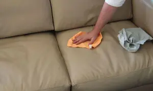 Mano che pulisce un divano in pelle beige con un panno arancione. Un altro panno grigio è appoggiato sul divano.