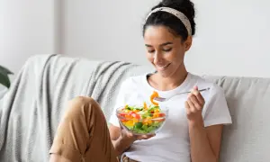 Donna sorridente mangia un'insalata colorata sul divano, godendosi un momento di relax e benessere.