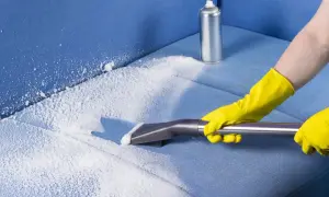 Mani con guanti gialli eseguono il lavaggio divano a secco con schiuma e aspiratore su tessuto blu.