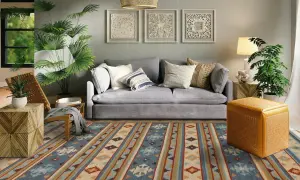 Tappeto 2 metri per 3 con motivi geometrici in blu, beige e arancione, che copre il pavimento di un soggiorno moderno con divano grigio e piante verdi.