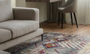 Un tappeto colorato sotto un divano beige, con una scrivania e una sedia sullo sfondo. Tappeto sotto divano si o no? La risposta è sì per aggiungere stile.