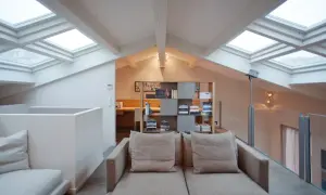 Accogliente salotto con soppalco, illuminato da lucernari. Divano beige, libreria moderna e scrivania in legno.
