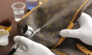 Mani guantate usano un aerografo per pulire la similpelle di una borsa monogrammata, mostrando un metodo per come pulire la similpelle.