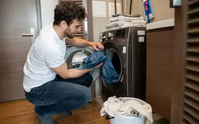 Uomo con maglietta bianca e jeans che carica la lavatrice, preoccupato per come eliminare le macchie da trasferimento di colore blu da un capo.
