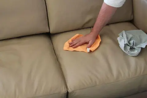 Mano che pulisce un divano in pelle beige con un panno arancione. Un altro panno grigio è appoggiato sul divano.