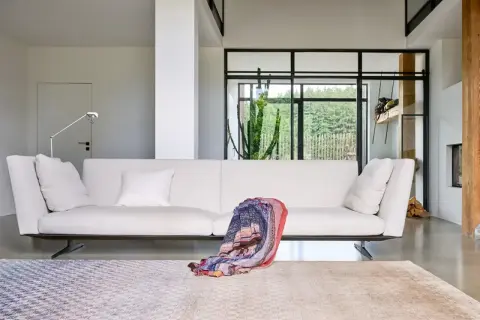 Divano bianco moderno, un plaid colorato appoggiato sopra, tappeti geometrici e un grande cactus dietro la vetrata. Design Jan Kath.