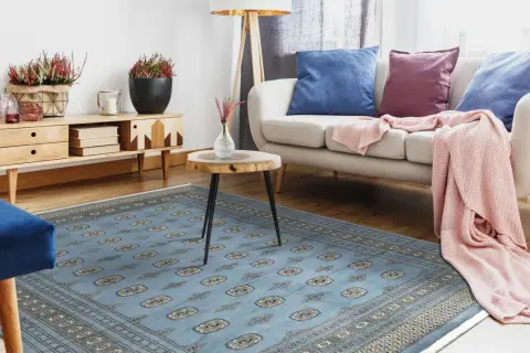 Un soggiorno moderno con un tappeto pakistano blu chiaro, divano beige, cuscini blu e viola, e una coperta rosa.