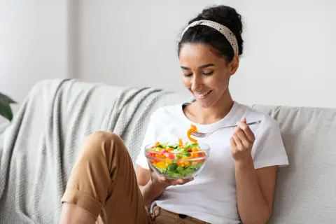 Donna sorridente mangia un'insalata colorata sul divano, godendosi un momento di relax e benessere.