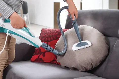 Come togliere macchie dal divano con un pulitore a vapore. La persona pulisce un cuscino del divano con un accessorio a spazzola.