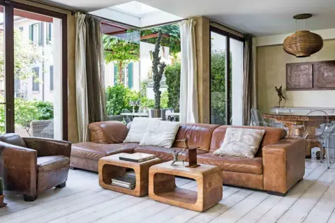 Soggiorno etnico chic con divano in pelle marrone, poltrona vintage e tavolini in legno grezzo. Ampie vetrate affacciano su un rigoglioso giardino.