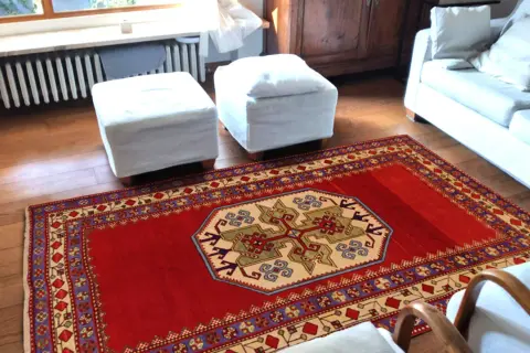 Un tappeto rosso con motivi geometrici, che evoca l'autenticità dei tappeti rustici, adorna il pavimento in legno di un soggiorno accogliente.