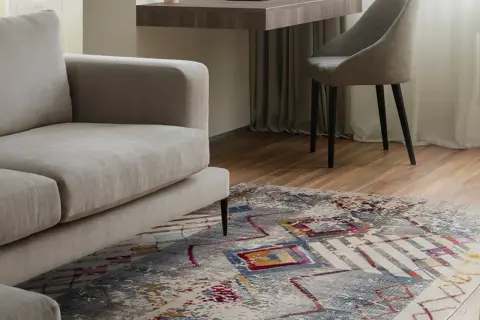 Un tappeto colorato sotto un divano beige, con una scrivania e una sedia sullo sfondo. Tappeto sotto divano si o no? La risposta è sì per aggiungere stile.
