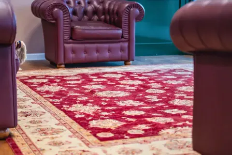 Un tappeto Yalameh rosso con motivi floreali chiari, adagiato su un pavimento in legno, accanto a poltrone in pelle bordeaux.
