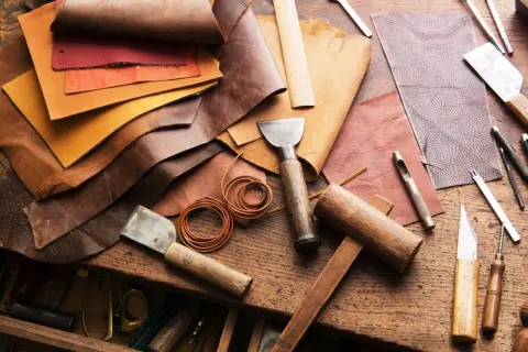 Pelle, colori e attrezzi da artigiano. Un tavolo da lavoro con vari tipi di pelle, strumenti per lavorarla come punzoni, coltelli e martelli.