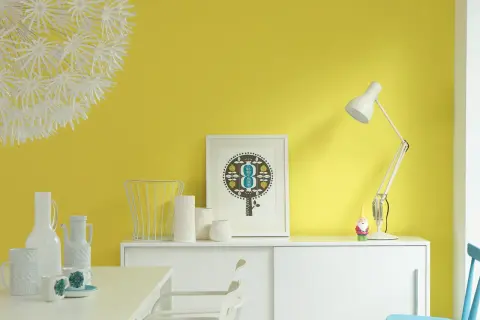Parete giallo dorato illumina un mobile bianco con vasi, una lampada e un quadro. Un piccolo gnomo sorveglia la scena.