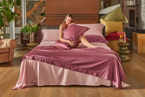 Una donna sorride su un letto con lenzuola rosa. La trapunta nuova va lavata prima di usarla.