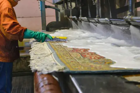 Un operaio lava un tappeto persiano con una spazzola, sotto getti d'acqua e schiuma. Il costo del lavaggio tappeto persiano è un investimento per preservarne la bellezza.