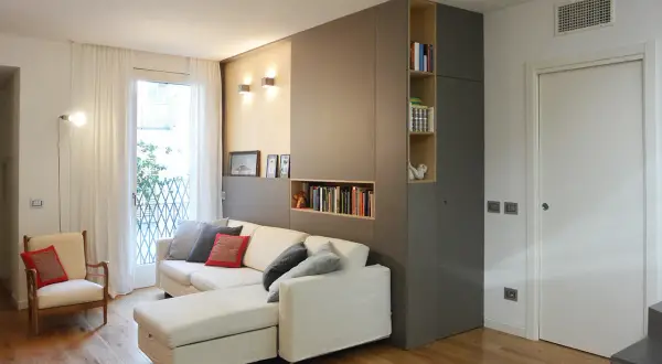 Divano bianco con cuscini grigi e rossi, accanto a una libreria a muro color un marrone chiaro.