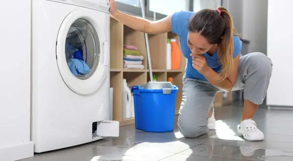 Donna preoccupata controlla la lavatrice che perde acqua da sotto. Sul pavimento bagnato, un secchio blu e uno straccio.