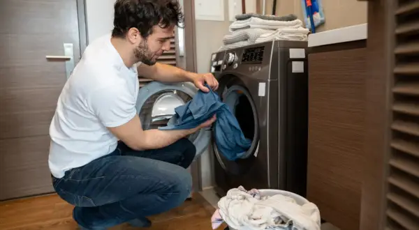 Uomo con maglietta bianca e jeans che carica la lavatrice, preoccupato per come eliminare le macchie da trasferimento di colore blu da un capo.