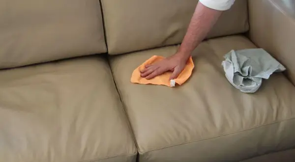 Mano che pulisce un divano in pelle beige con un panno arancione. Un altro panno grigio è appoggiato sul divano.