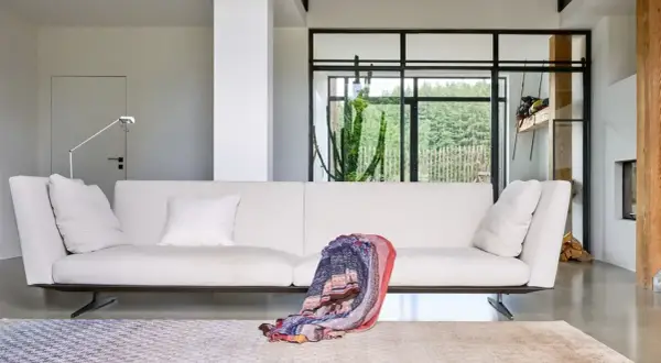 Divano bianco moderno, un plaid colorato appoggiato sopra, tappeti geometrici e un grande cactus dietro la vetrata. Design Jan Kath.