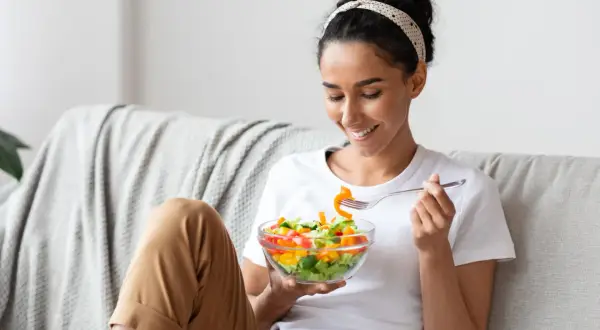 Donna sorridente mangia un'insalata colorata sul divano, godendosi un momento di relax e benessere.