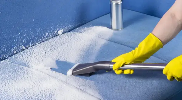 Person in yellow gloves using a vacuum attachment to clean a blue fabric sofa with white foam.