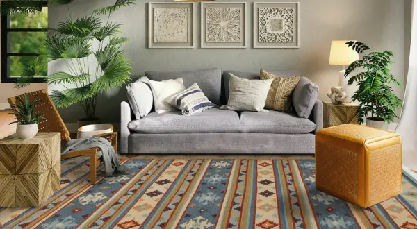 Tappeto 2 metri per 3 con motivi geometrici in blu, beige e arancione, che copre il pavimento di un soggiorno moderno con divano grigio e piante verdi.