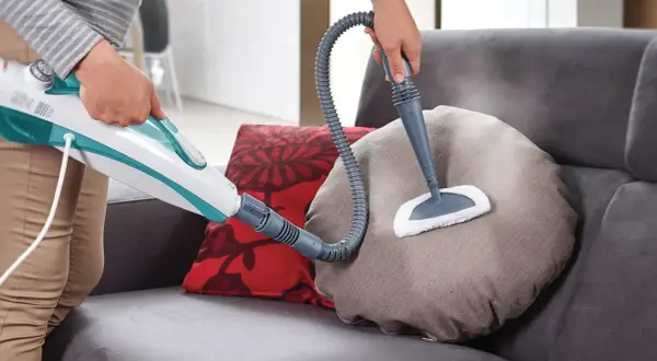 Come togliere macchie dal divano con un pulitore a vapore. La persona pulisce un cuscino del divano con un accessorio a spazzola.