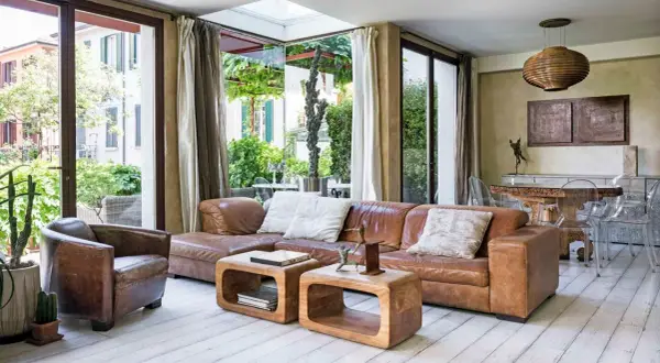 Soggiorno etnico chic con divano in pelle marrone, poltrona vintage e tavolini in legno grezzo. Ampie vetrate affacciano su un rigoglioso giardino.