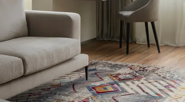 Un tappeto colorato sotto un divano beige, con una scrivania e una sedia sullo sfondo. Tappeto sotto divano si o no? La risposta è sì per aggiungere stile.
