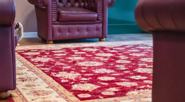 Un tappeto Yalameh rosso con motivi floreali chiari, adagiato su un pavimento in legno, accanto a poltrone in pelle bordeaux.