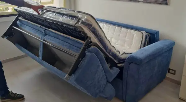 Apertura divano letto: un braccio solleva il meccanismo interno, rivelando il materasso ancora imballato.