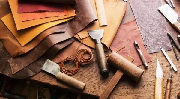 Pelle, colori e attrezzi da artigiano. Un tavolo da lavoro con vari tipi di pelle, strumenti per lavorarla come punzoni, coltelli e martelli.