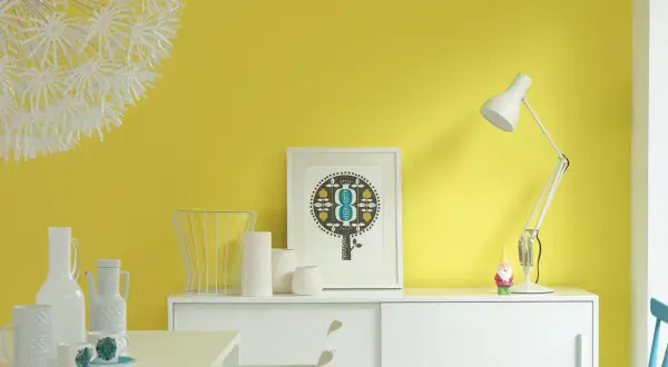 Parete giallo dorato illumina un mobile bianco con vasi, una lampada e un quadro. Un piccolo gnomo sorveglia la scena.