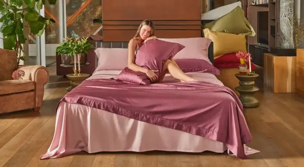 Una donna sorride su un letto con lenzuola rosa. La trapunta nuova va lavata prima di usarla.