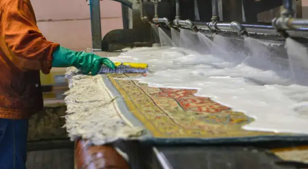 Un operaio lava un tappeto persiano con una spazzola, sotto getti d'acqua e schiuma. Il costo del lavaggio tappeto persiano è un investimento per preservarne la bellezza.