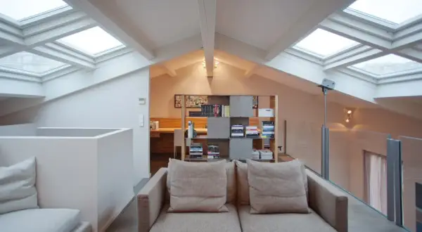 Accogliente salotto con soppalco, illuminato da lucernari. Divano beige, libreria moderna e scrivania in legno.