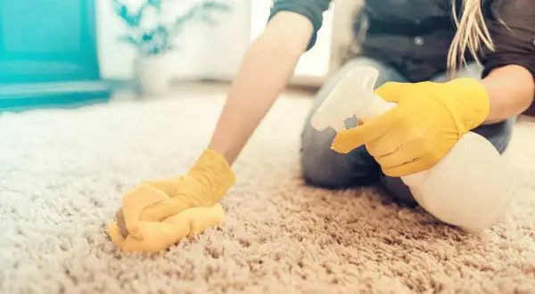 Mani con guanti gialli spruzzano detergente su un tappeto chiaro, mostrando come pulire un tappeto a secco con un panno.