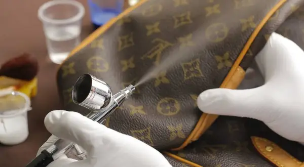 Mani guantate usano un aerografo per pulire la similpelle di una borsa monogrammata, mostrando un metodo per come pulire la similpelle.