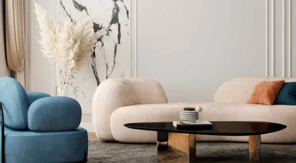 Un accogliente soggiorno con un divano beige, pareti chiare e una poltrona blu. Un tavolino nero lucido con base dorata completa l'arredamento.