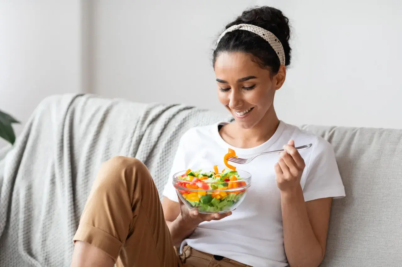 Donna sorridente mangia un'insalata colorata sul divano, godendosi un momento di relax e benessere.