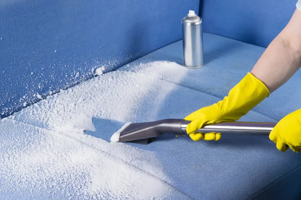 Person in yellow gloves using a vacuum attachment to clean a blue fabric sofa with white foam.