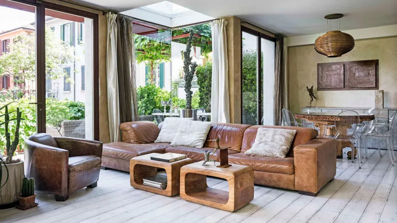Soggiorno etnico chic con divano in pelle marrone, poltrona vintage e tavolini in legno grezzo. Ampie vetrate affacciano su un rigoglioso giardino.