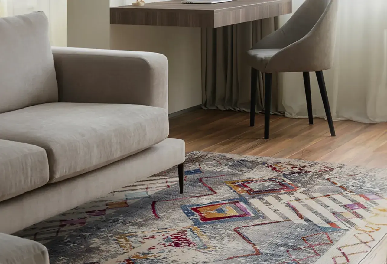 Un tappeto colorato sotto un divano beige, con una scrivania e una sedia sullo sfondo. Tappeto sotto divano si o no? La risposta è sì per aggiungere stile.