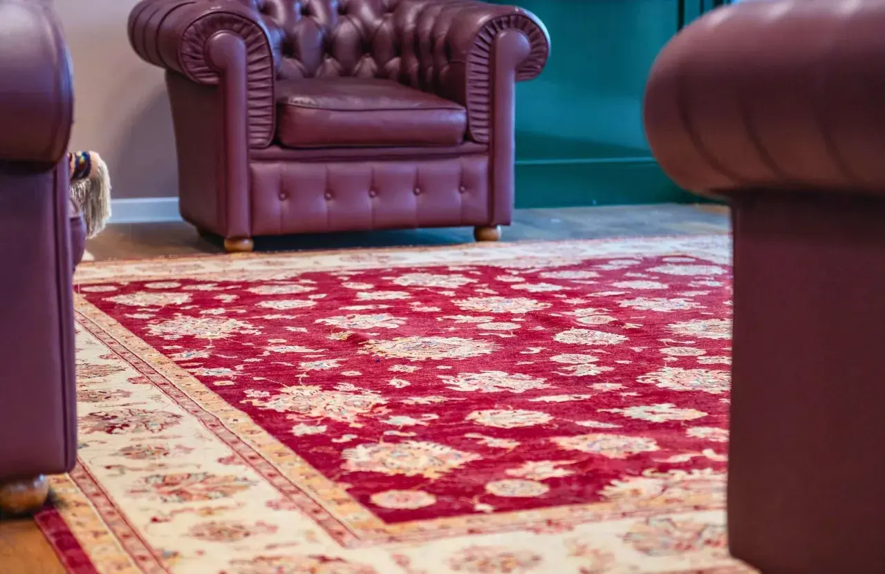 Un tappeto Yalameh rosso con motivi floreali chiari, adagiato su un pavimento in legno, accanto a poltrone in pelle bordeaux.