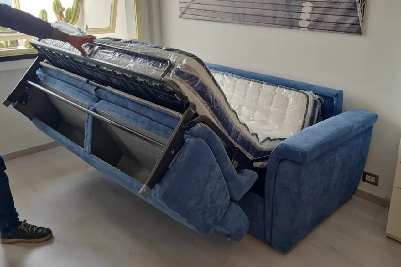 Apertura divano letto: un braccio solleva il meccanismo interno, rivelando il materasso ancora imballato.